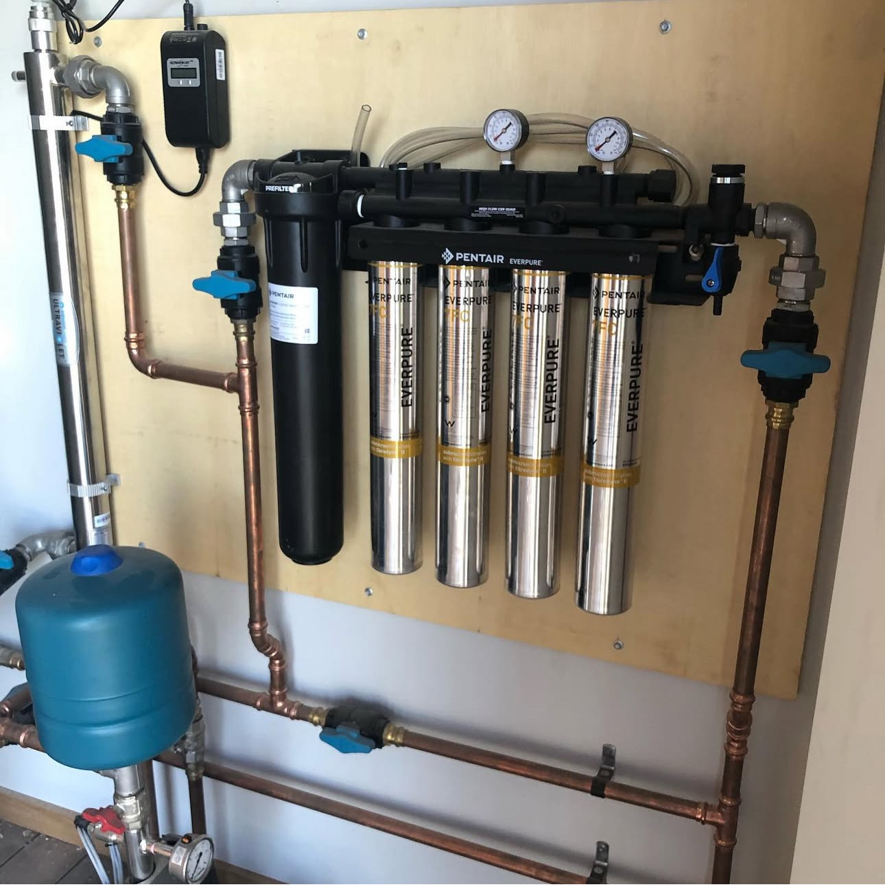 A whole house water treatment installation on a ply backboard: a four cartridge filtration manifold, prefilter, UV steriliser and pressure vessel, hard plumbed in copper
