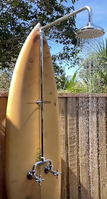 A custom outdoor shower plumbed against a surfboard mounted on a timber fence, with chrome cross handles and a rain head